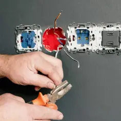 Dłonie trzymające kombinerki przy przewodach elektrycznych podłączanych do gniazdka.
