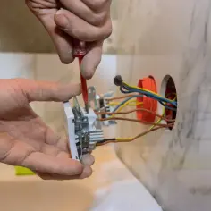 Ręce montują gniazdo elektryczne, podłączając przewody. Widać śrubokręt i kolorowe kable wychodzące z puszki.