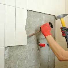 Ręce w rękawiczkach usuwają stare płytki z kleju za pomocą dłuta i młotka.