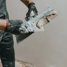 Ręce w rękawiczkach nakładają gipsową masę na pacę, przygotowując się do pracy. Dowiedz się, jak wygładzić tynk gipsowy.