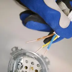 Ręka w rękawicy przygotowuje przewody do budowy gniazdka elektrycznego. Widoczne gołe końcówki kabli: brązowy, niebieski i żółto-zielony.