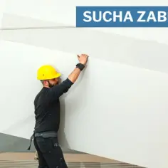 Budowlaniec w żółtym kasku przenosi dużą płytę gipsową. W tle widać stos płyt i podnośnik. Płyta gipsowa wymiar jest kluczowy dla prac.