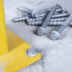 Montaż słupków ogrodzeniowych na kotwach. Metalowe śruby z gwintem i łbem sześciokątnym leżą obok żółtej podstawy.