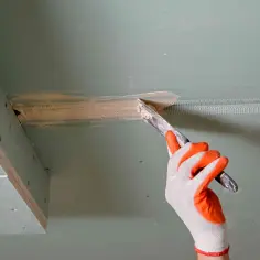 Ręka w rękawicy nakłada masę szpachlową na taśmę zbrojącą, przygotowując do klejenia płyt gk na piankę.