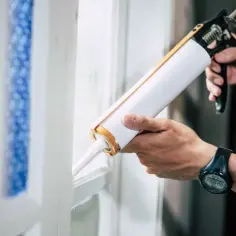 Dłoń trzymająca pistolet do silikonu i aplikująca go na ramę okna.