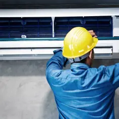 Technik w żółtym kasku wykonuje montaż klimatyzacji krok po kroku, przykręcając element śrubokrętem.