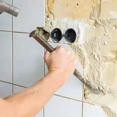 Ręka z młotkiem i dłutem usuwa stare płytki i klej ze ściany, odsłaniając puszki elektryczne.