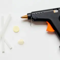 Czarny pistolet do klejenia z pomarańczowym spustem, obok kilka pałeczek kleju, okrągły element i muszelka.