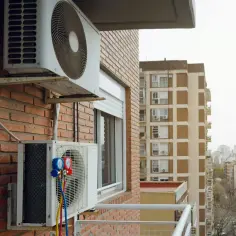 Montaż klimatyzacji w mieszkaniu. Dwa urządzenia wiszą na ceglanej ścianie, jedno z manometrami. W tle widać miejską zabudowę.