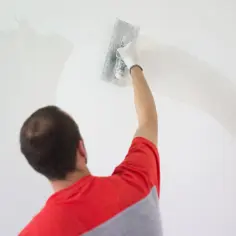 Mężczyzna w czerwonej bluzie nakłada gładź szpachlową na ścianę. Czas, ile schnie gładź szpachlowa, zależy od warunków.