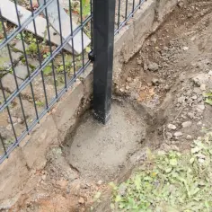 Trwa osadzanie słupków ogrodzeniowych. Widać wykopany dół z betonem, w którym umieszczono czarny słup ogrodzeniowy.