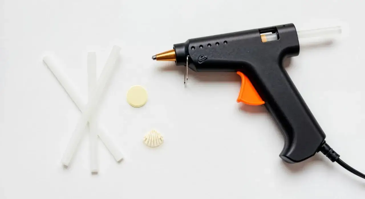 Czarny pistolet do klejenia z pomarańczowym spustem, obok pałeczki kleju, muszelka i żółty krążek.