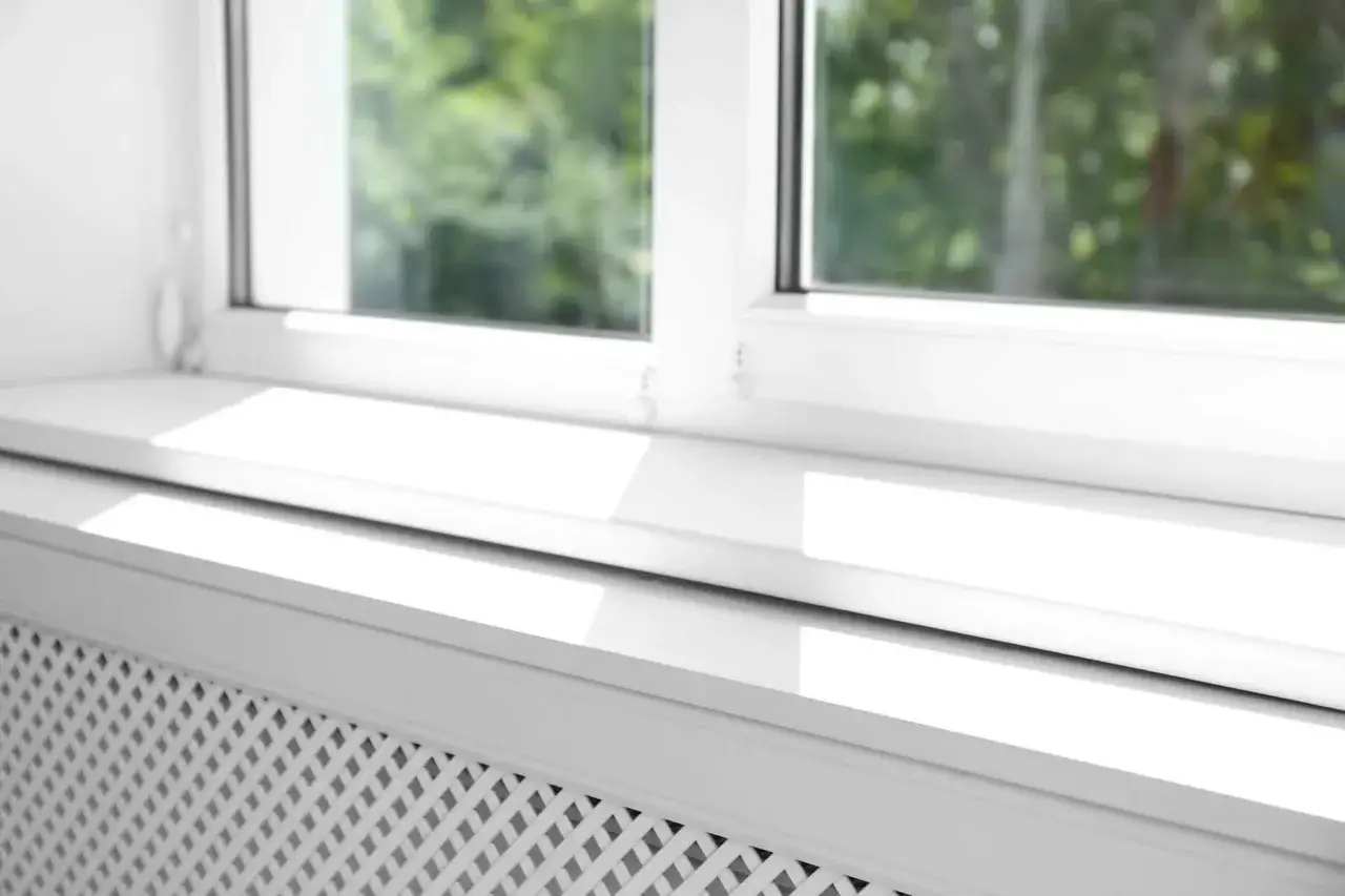 Biały parapet zlicowany ze ścianą, z widocznym kratkowanym grzejnikiem pod nim. Słońce rzuca jasne refleksy na gładką powierzchnię.