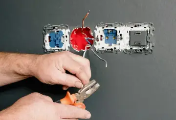 Dłonie trzymające kombinerki przy przewodach elektrycznych podłączanych do gniazdka.