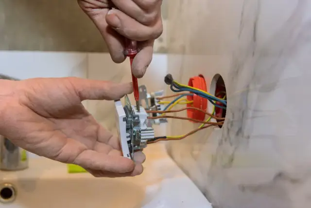 Ręce montują gniazdo elektryczne, podłączając przewody. Widać śrubokręt i kolorowe kable wychodzące z puszki.