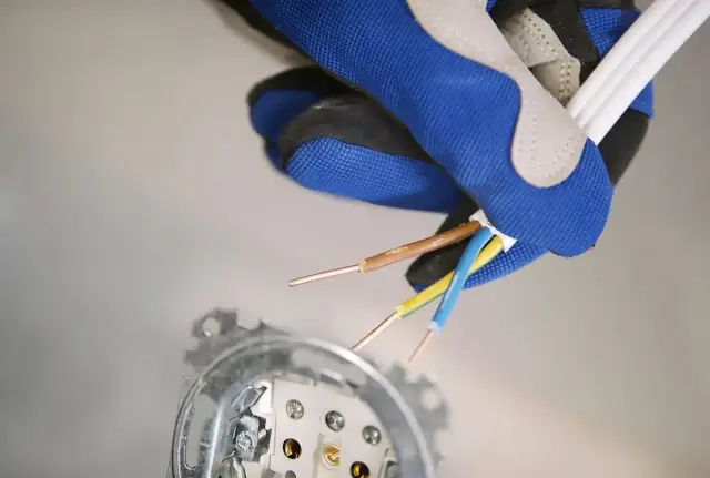 Ręka w rękawicy przygotowuje przewody do budowy gniazdka elektrycznego. Widoczne gołe końcówki kabli: brązowy, niebieski i żółto-zielony.