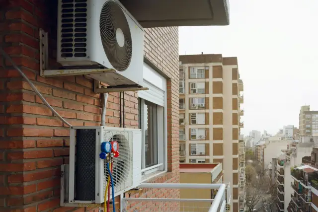 Montaż klimatyzacji w mieszkaniu. Dwa urządzenia wiszą na ceglanej ścianie, jedno z manometrami. W tle widać miejską zabudowę.